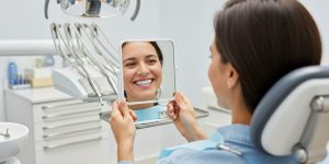 A patient in a dental chair holds a mirror and smiles at her bright, newly whitened teeth, enjoying the smile aesthetics benefits of teeth whitening treatment.