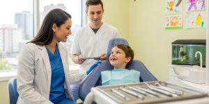 A friendly female dentist providing dental services you can expect from a family dentist to a young boy in a dental chair, with a male assistant standing nearby.